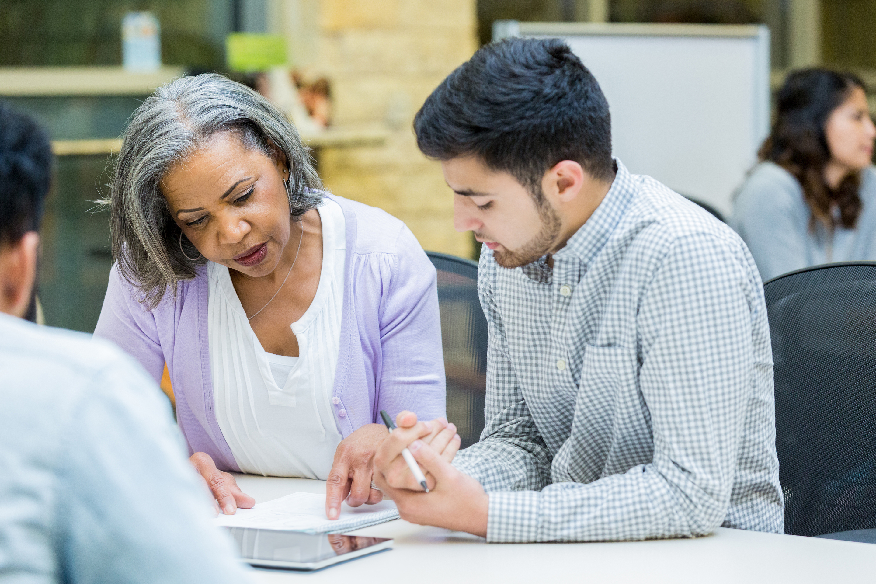 Diverse business people review information during weekly staff meeting. An African American woman points to something on a note pad. A Middle Eastern man is attentively listening to the woman