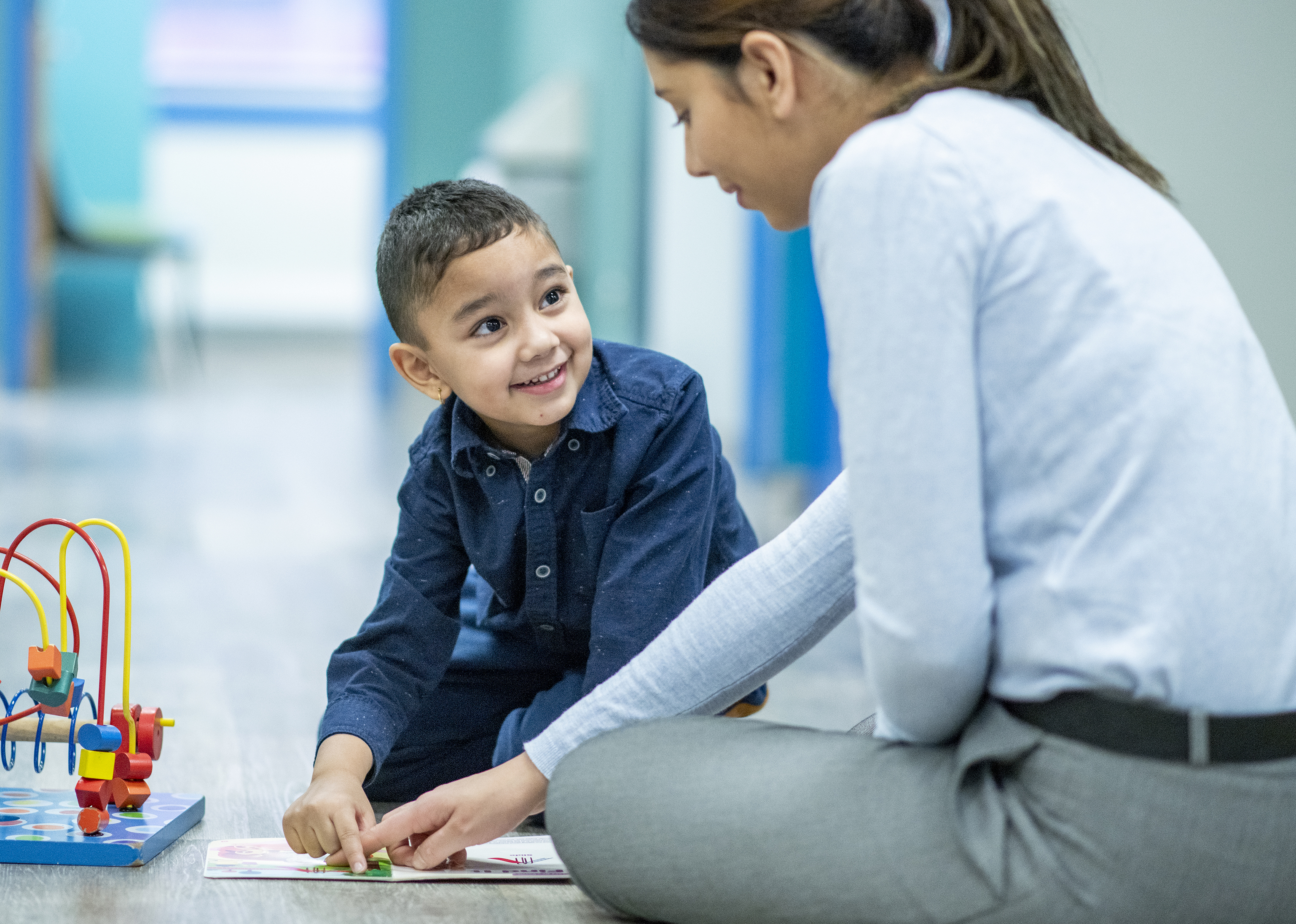 A young female, mixed race, Occupational Therapist sits cross-legged on the floor of a hospital beside her young patient.  She is dressed semi-casually in dress pants and a button-up blouse, and is engaging the young boy with an interactive bk she has laying out on the floor between them.  The young boy is kneeling on the floor in a button-up shirt as he works with the therapist.