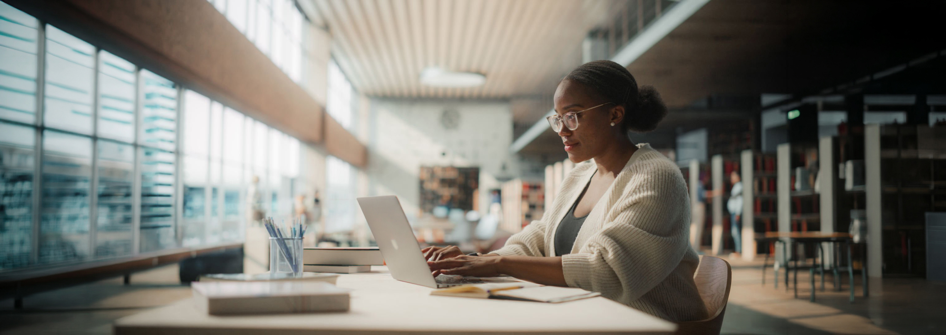 Dedicated African American Female Student Engaged in Online Learning at a Modern Library. Young Woman Using Laptop for Research, Surrounded by Books and Academic Environment.