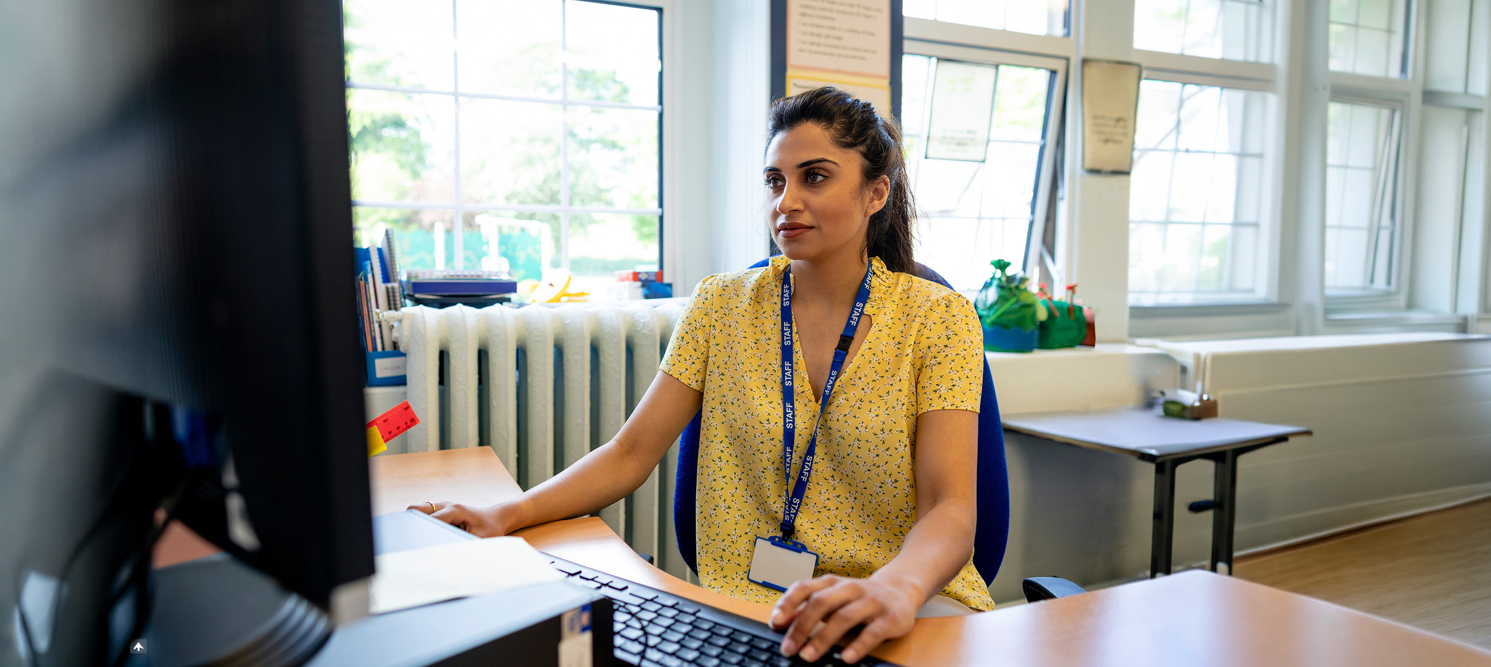 A close up front view of a young teacher sitting at her desk in her classroom. She is checking her lesson plans on her desktop PC. She is working in a school in Hexham in the North East of England.